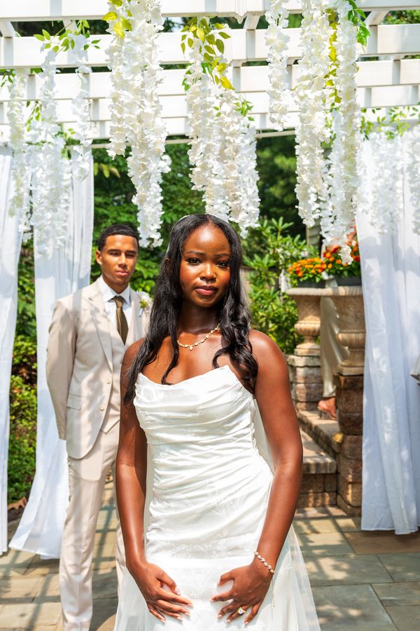 Bride in a white strapless gown looks into the camera under hanging white wisteria, the groom standing behind her