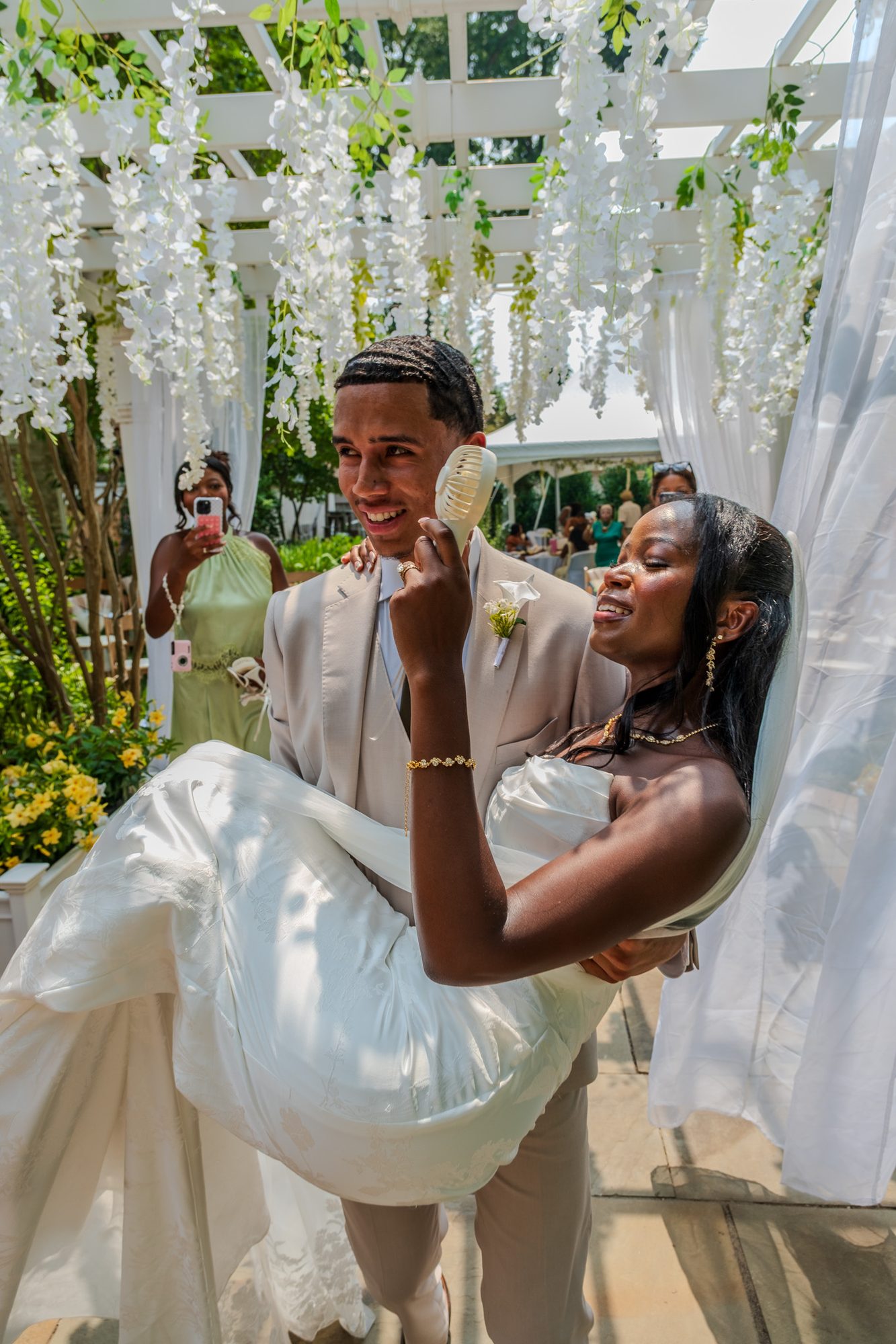 A groom carries his bride under a pergola of white wisteria while she laughs and holds a handheld fan